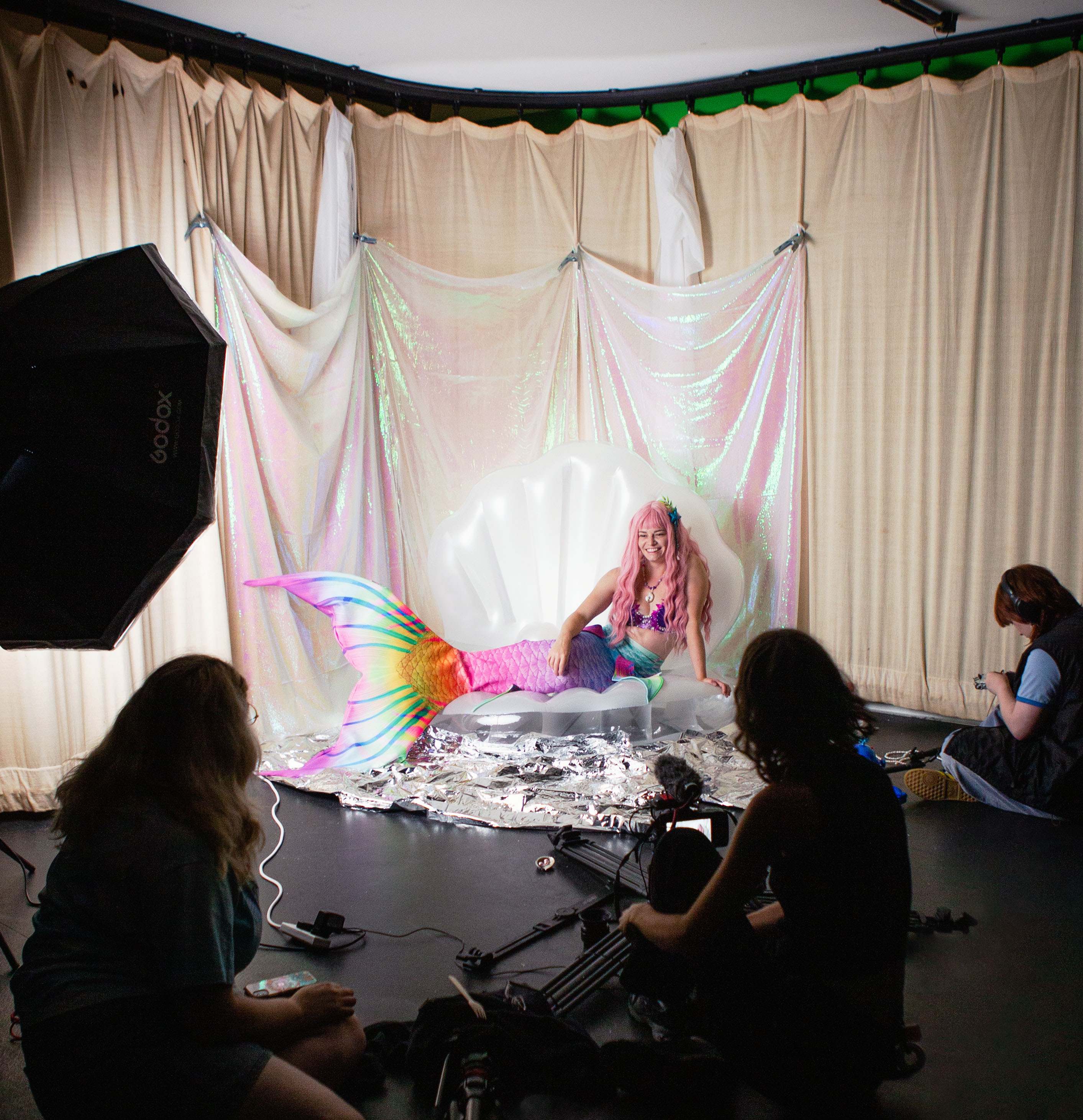 Kaia the mermaid sits in an enclosed studio space that has been fully dressed for a mermaid with a sparkling backdrop, bubbles, an inflatable shell, and Kaia in a wig, mermaid tail, and seashell bra. Sitting around her are the crew composed of camera, sound, and director.
